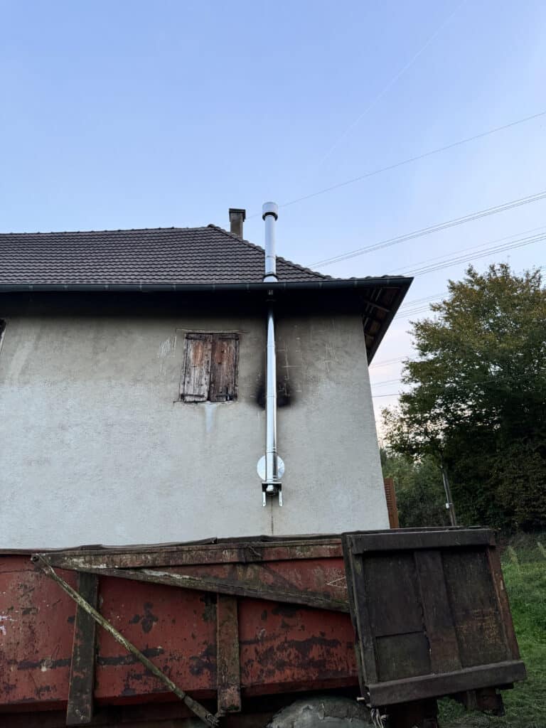 Maison à façade claire et cheminée extérieure en acier tachée de suie noire. Une vieille remorque agricole rouillée rouge occupe le premier plan.