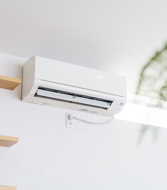 Climatiseur split system blanc fixé au mur, branché sur prise, près d'escaliers en bois et d'une fenêtre.