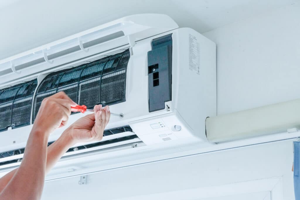 Mains d'un technicien nettoyant ou réparant un climatiseur mural blanc avec un tournevis, filtres encrassés visibles.