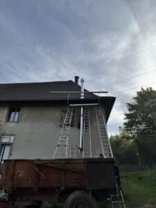 Nouvelle cheminée métallique moderne sur façade de maison en rénovation, avec échafaudage d'échelles. Un ancien camion rouillé est visible.