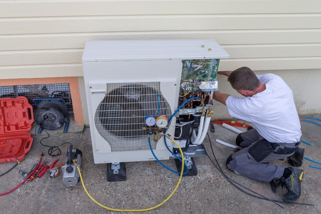 Technicien CVC installe une unité de climatisation extérieure, manomètres connectés. Outils de réparation au sol.