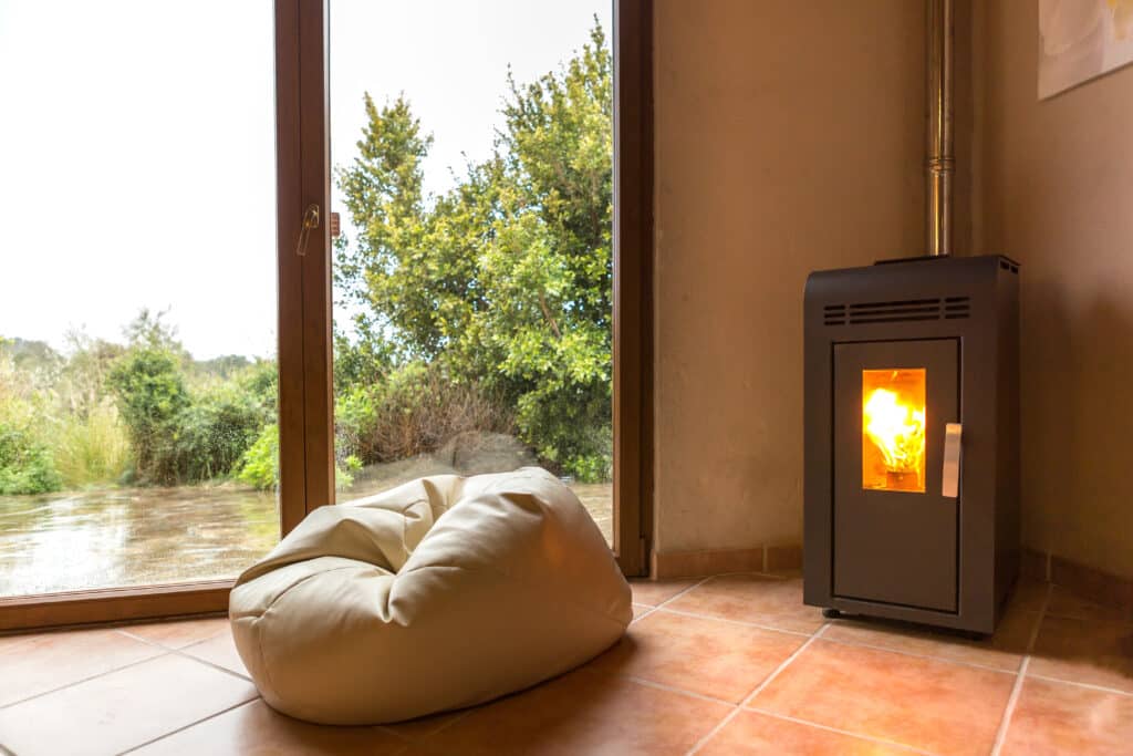 Intérieur douillet avec poêle à granulés anthracite aux flammes vives et pouf beige près d'une grande fenêtre donnant sur un paysage verdoyant.