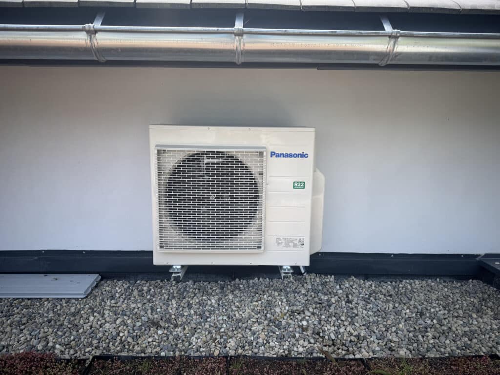 Unité extérieure de climatisation Panasonic R32, couleur crème, installée sur un toit en gravier sous une gouttière métallique.