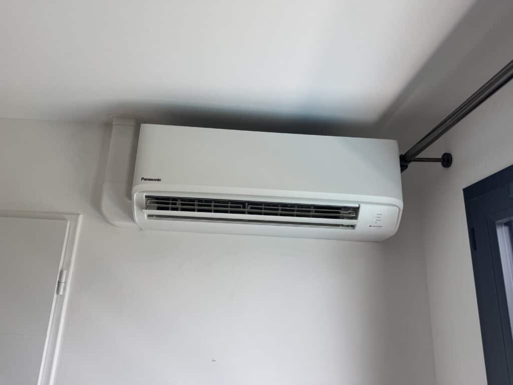 Climatiseur Panasonic mural blanc fixé sur un mur clair, avec les indicateurs POWER, TIMER et nanoeX visibles. Un conduit et une tringle noire sont aussi présents.