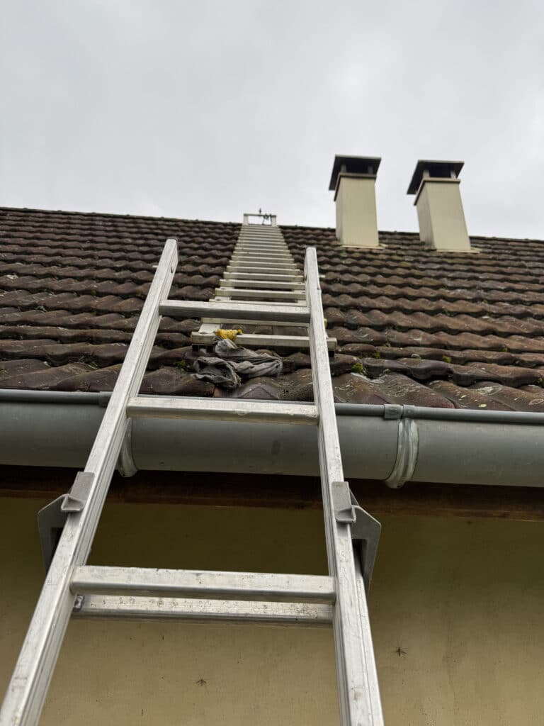 Échelle en aluminium installée sur un toit de tuiles anciennes, avec deux cheminées et gouttière visibles, sous ciel nuageux.