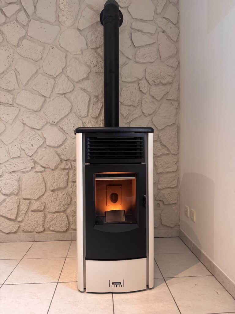 Poêle à granulés Superior blanc et noir allumé, cheminée avec conduit noir devant un mur en pierre claire et carrelage beige.