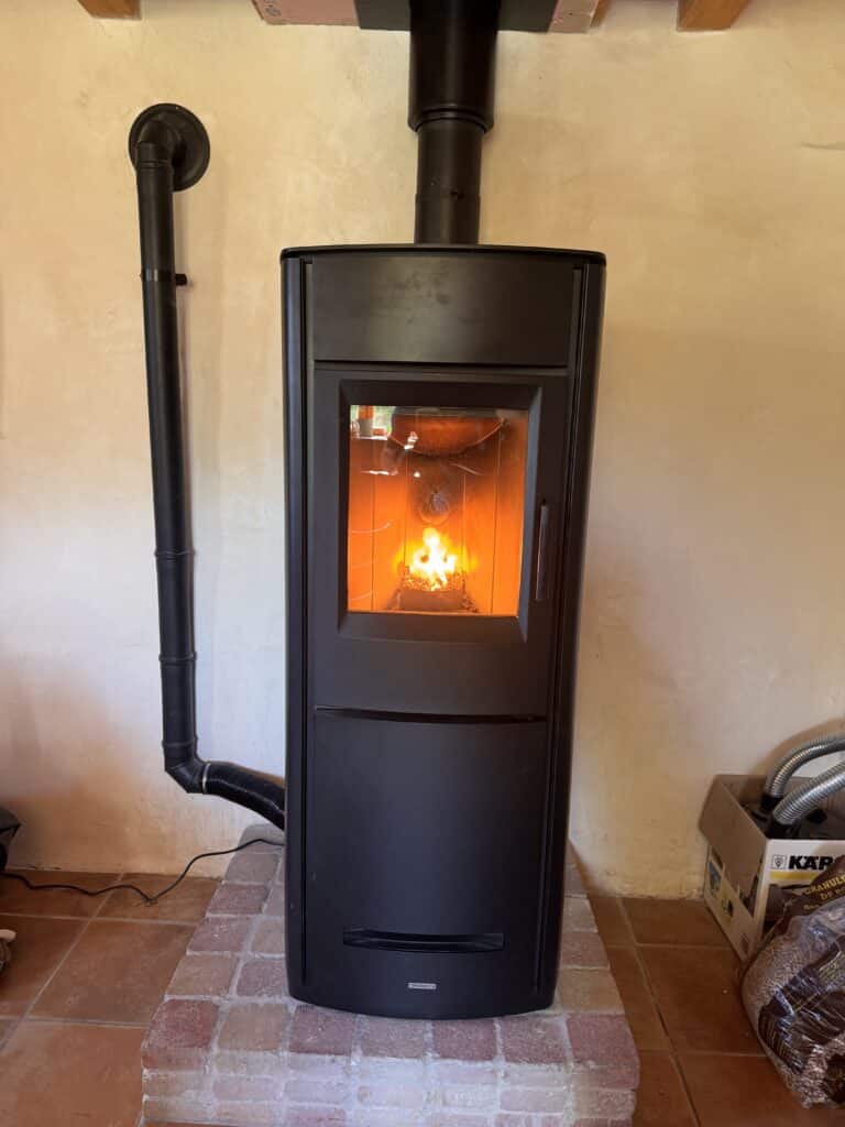 Poêle à granulés Piazzetta noir en fonctionnement, flammes visibles, raccordé à un conduit d'évacuation noir mural, sur socle en briques.