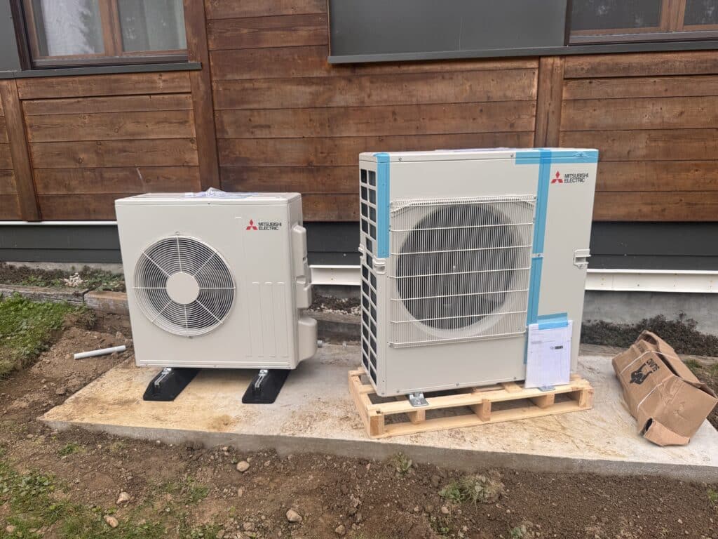 Deux unités extérieures de thermopompes Mitsubishi Electric neuves, installées sur une dalle de béton devant un revêtement en bois brun.